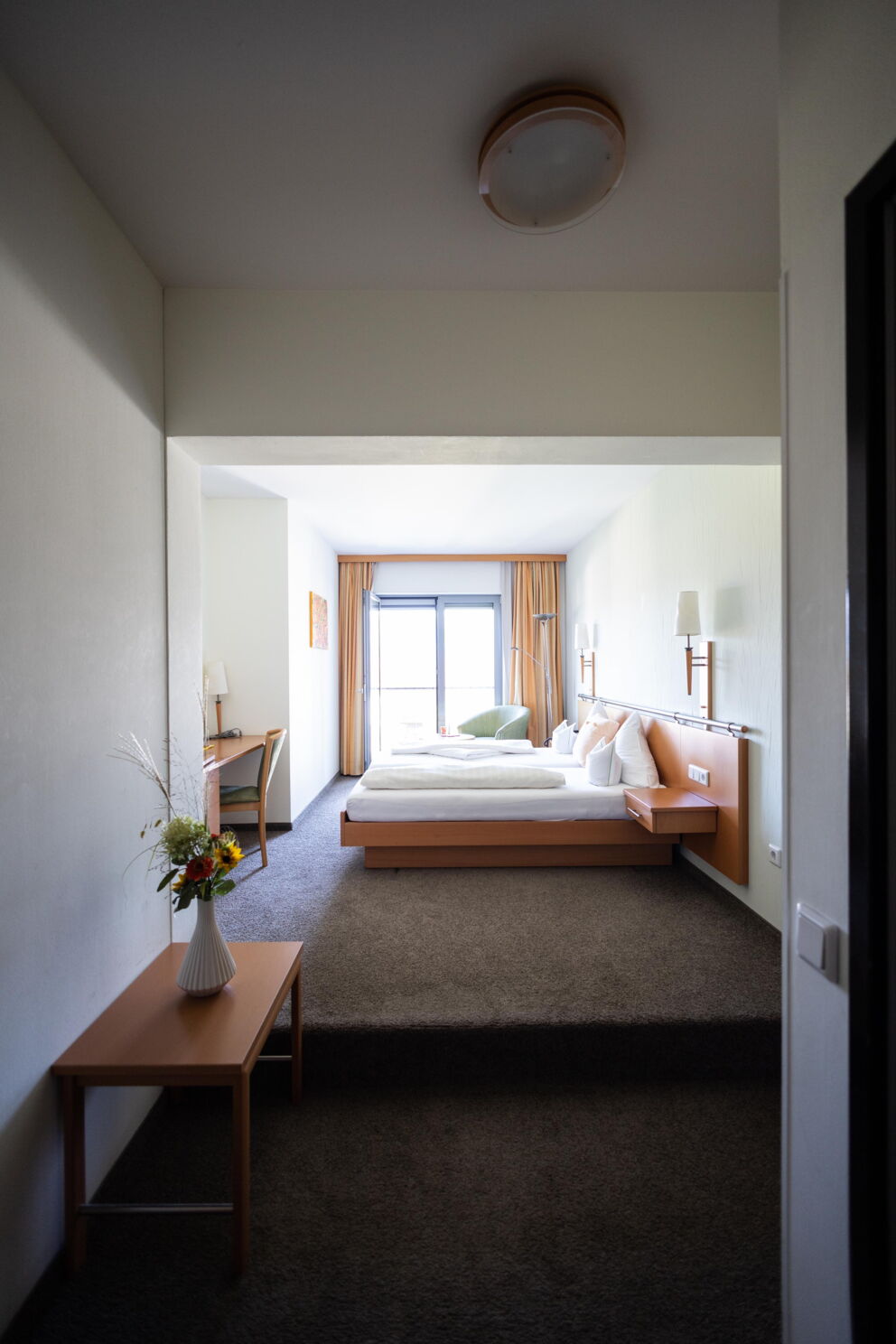 Helles Zimmer mit großem Fenster, Doppelbett und Tisch mit Blumenvasen.