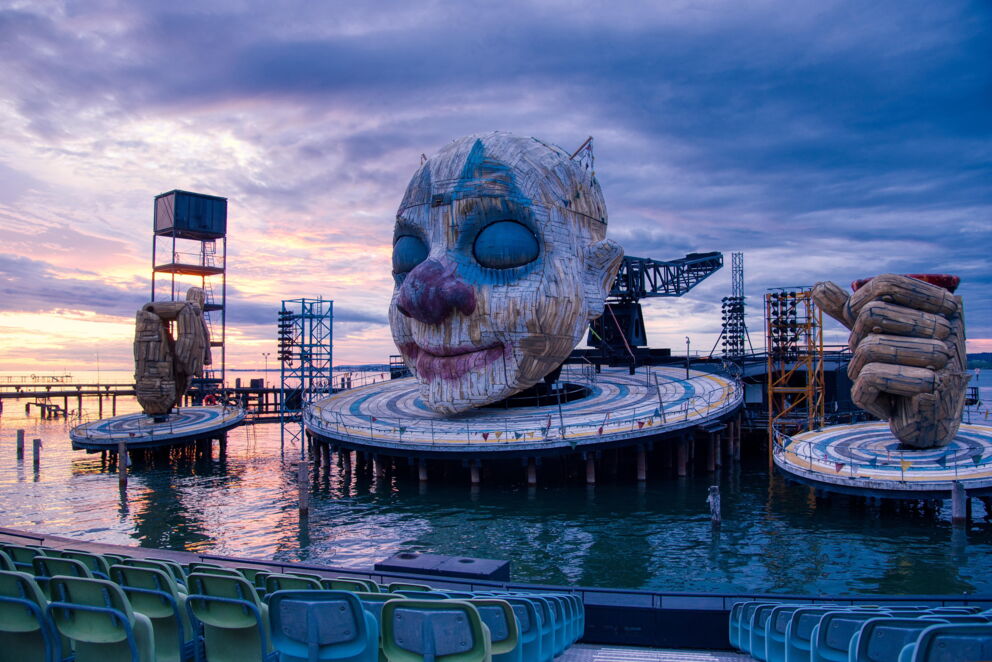 Large sculpture of a face and hands on a lake stage at sunset.