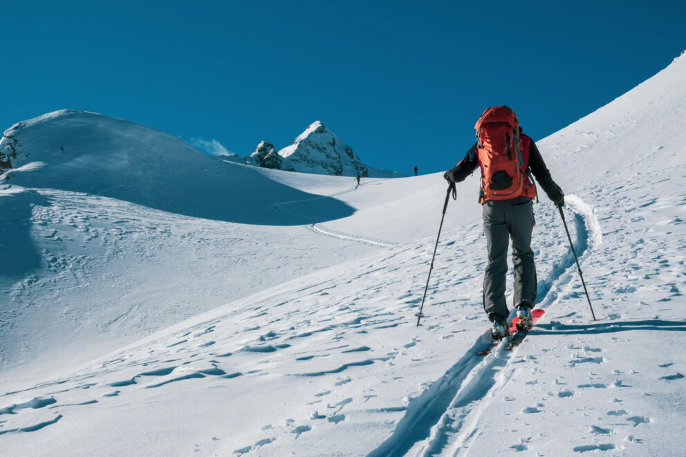 Skitourengeher auf schneebedecktem Hang unter klarem, blauem Himmel in alpiner Kulisse.
