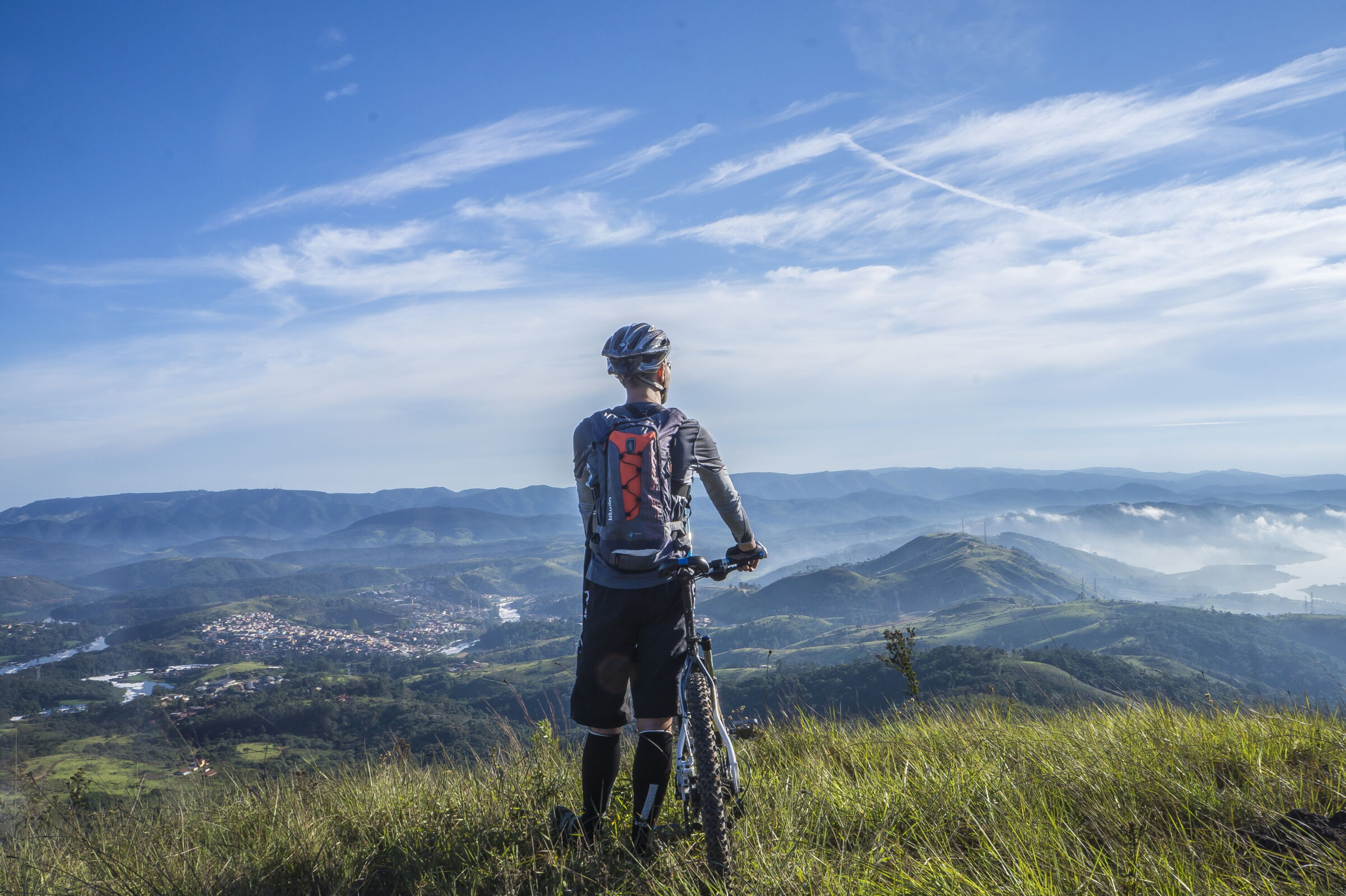 Mountainbiker steht auf Hügel und blickt auf weite Berglandschaft unter blauem Himmel.