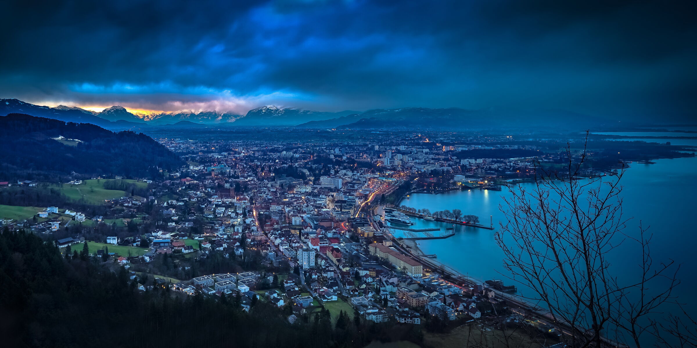 Stadtansicht bei Dämmerung mit Bergen und Wasser im Hintergrund unter bewölktem Himmel.