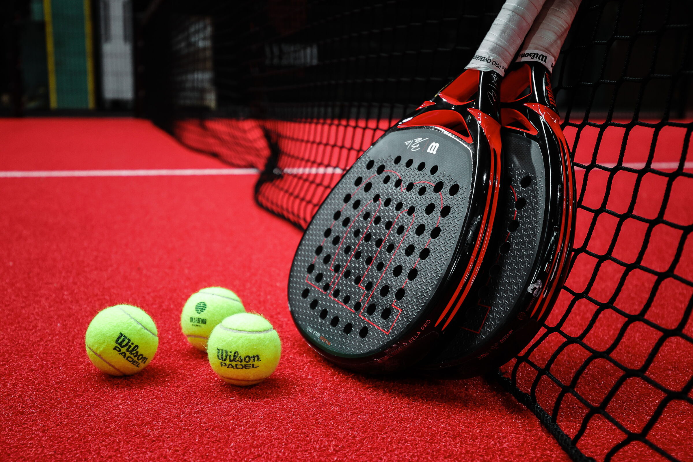 Zwei Padel-Schläger lehnen an einem Netz mit drei gelben Bällen auf rotem Boden.