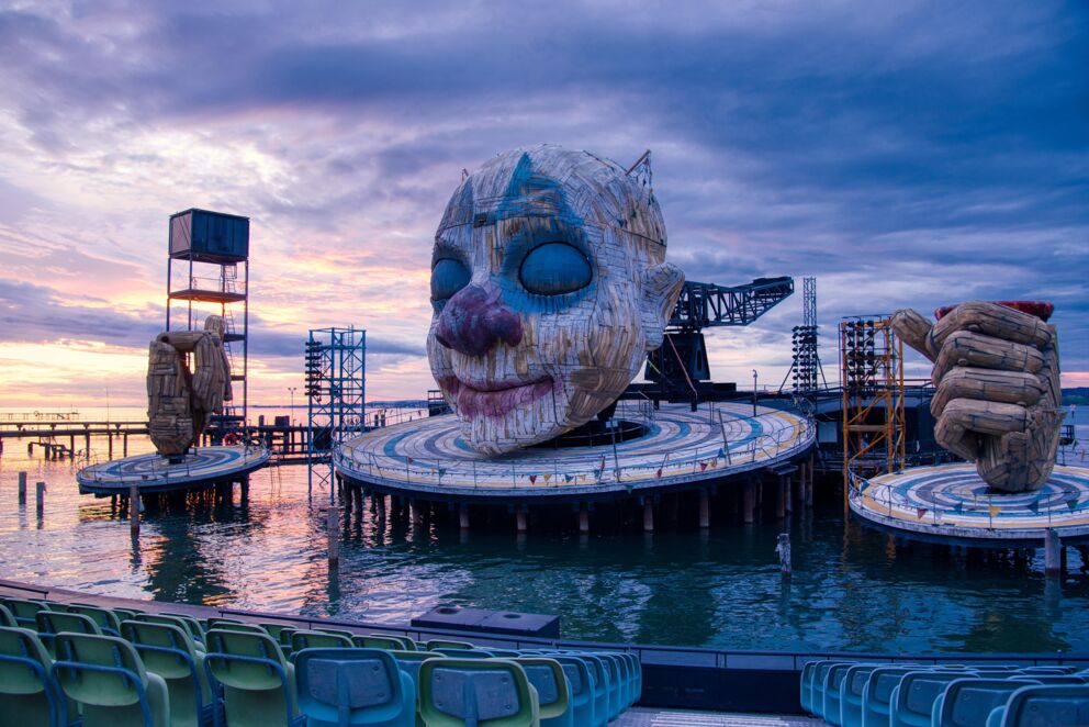 Large sculpture of a face and hands on a lake stage at sunset.