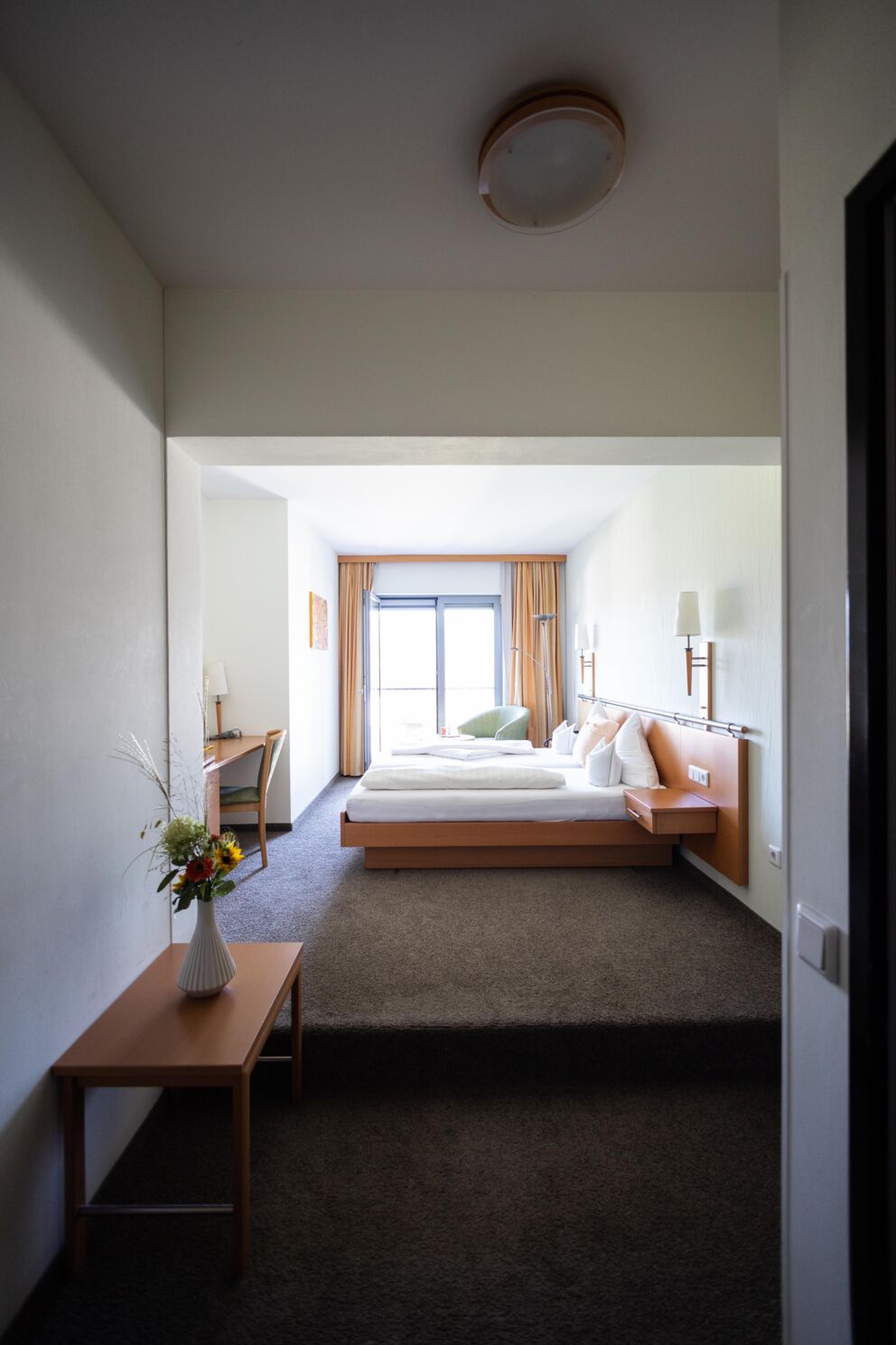 Helles Zimmer mit großem Fenster, Doppelbett und Tisch mit Blumenvasen.