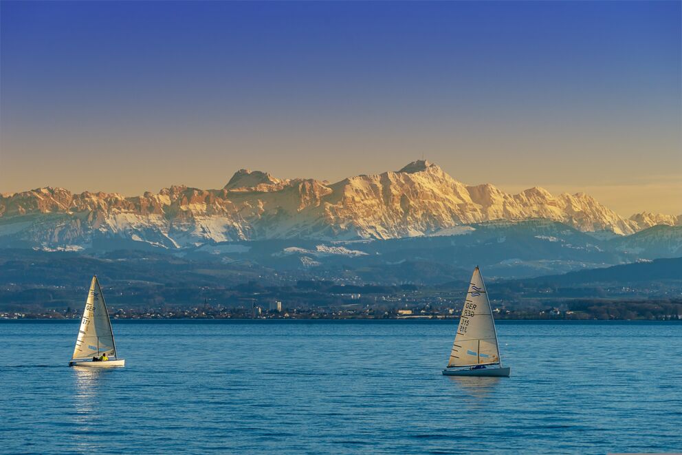 Two sailing boats on a lake with snow-capped mountains in the background.