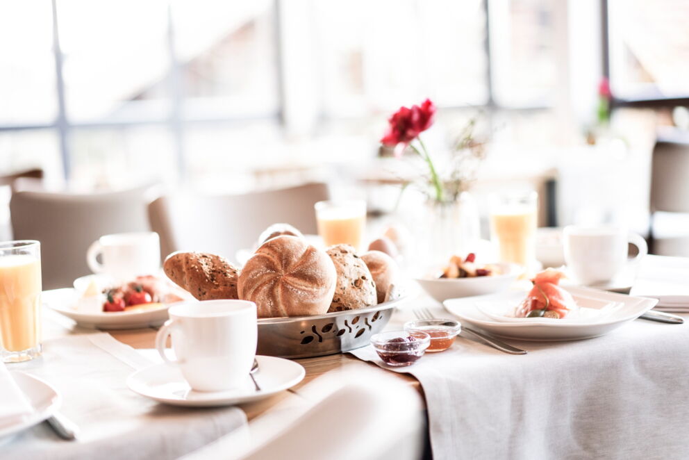 Frühstückstisch mit Brötchen, Kaffee, Saft und Blumen in heller Umgebung.
