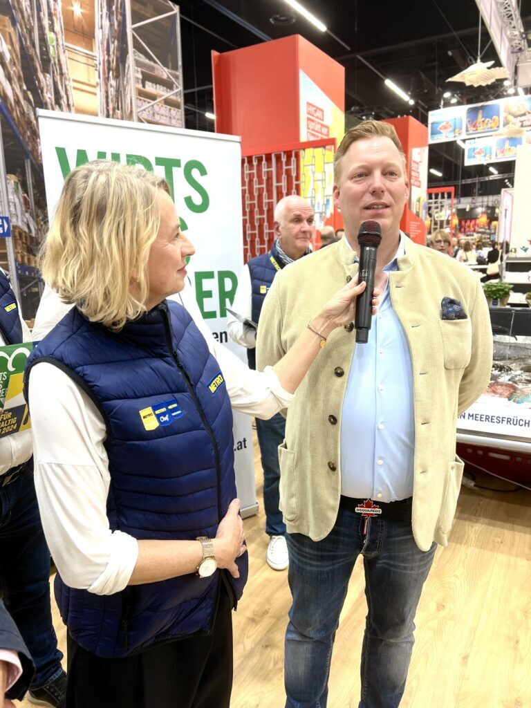 Zwei Personen im Interview bei einer Messeveranstaltung mit lebendigem Hintergrund.