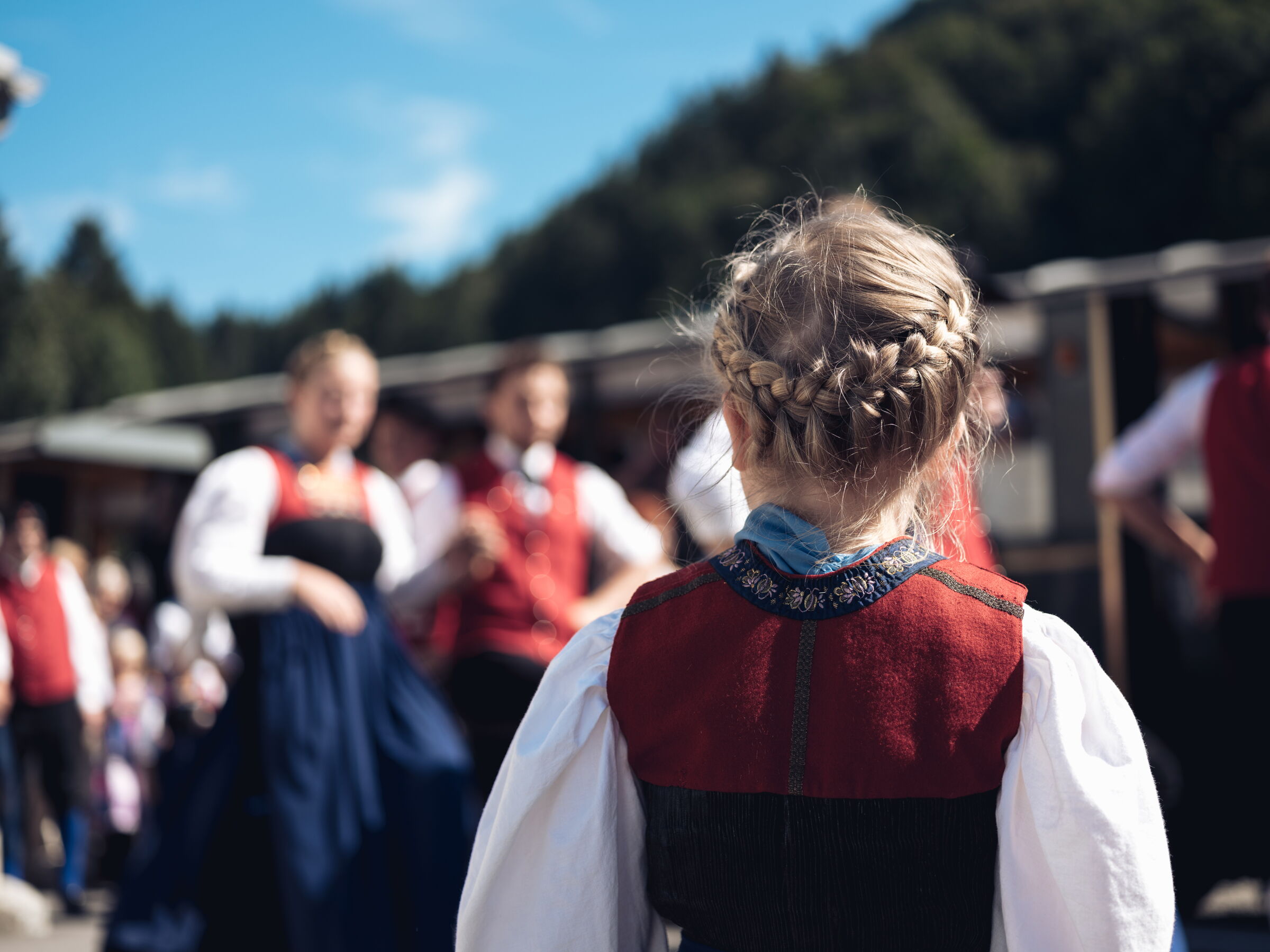 Mädchen in traditioneller Tracht beobachtet tanzende Gruppe im Freien bei sonnigem Wetter.