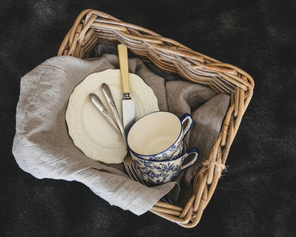 Woven basket with plates, cutlery, cups on linen cloth. © debby hudson by unsplash