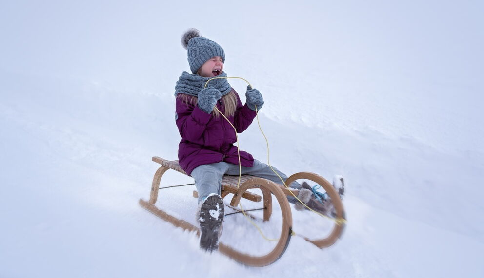Kind mit Winterkleidung rodelt freudig auf einem Holzschlitten durch verschneite Landschaft.