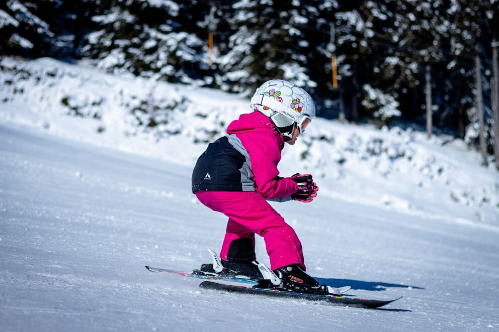 Kind in pinker Skijacke fährt auf schneebedeckter Piste im Sonnenlicht.