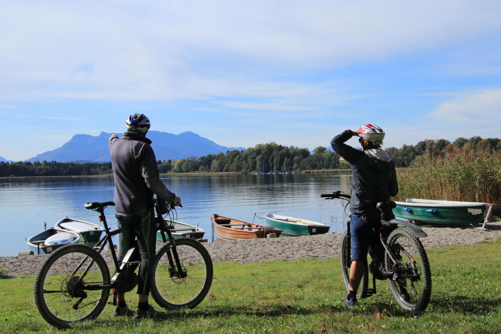 Two cyclists in front of a lake with boats and mountain views in the background.