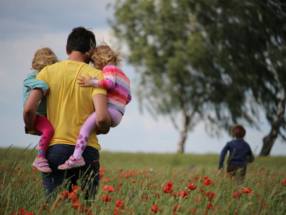 Vater trägt zwei Kinder durch eine blühende Wiese, weiteres Kind läuft voraus.