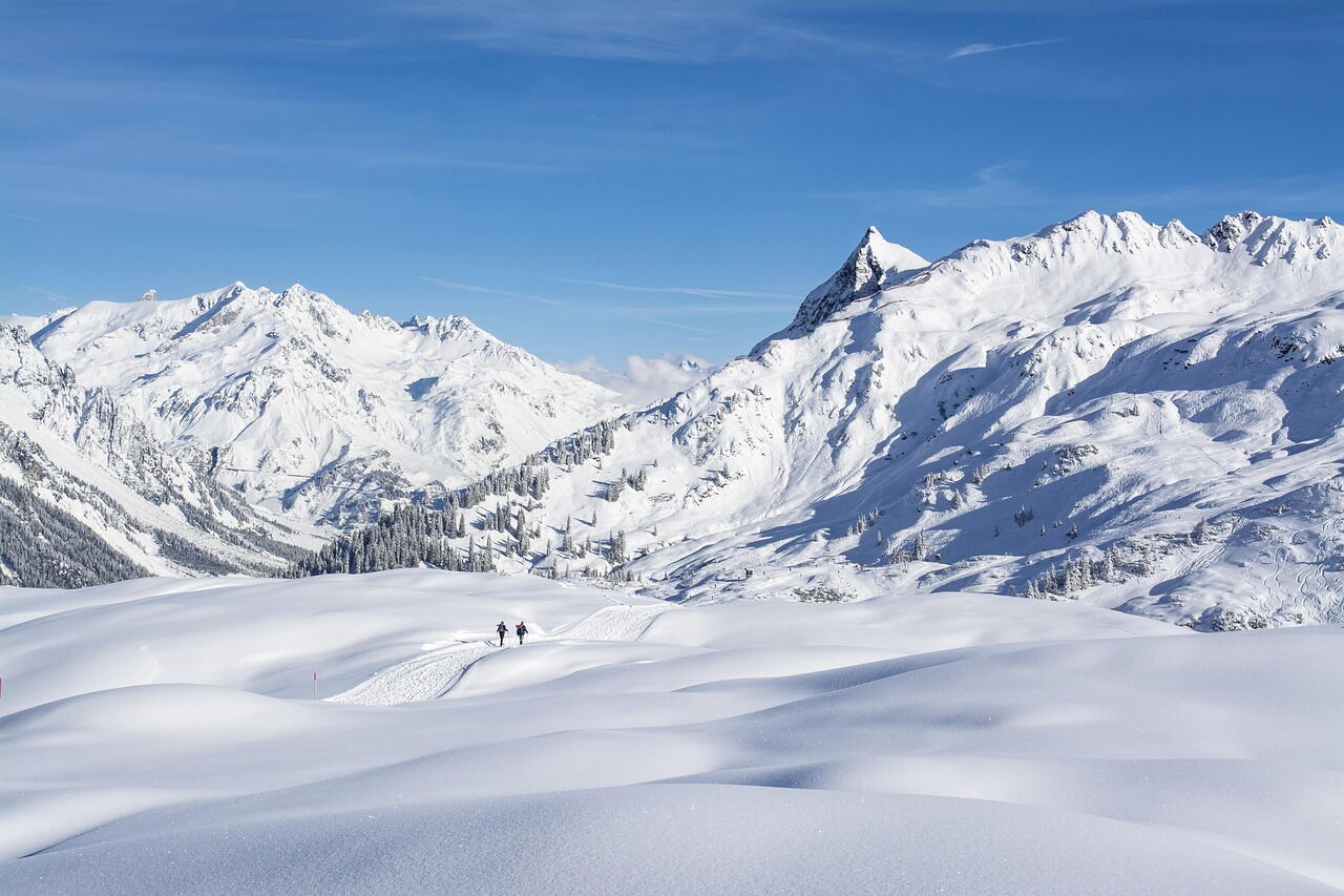Two hikers in a snow-covered, majestic Alpine landscape under a clear blue sky.