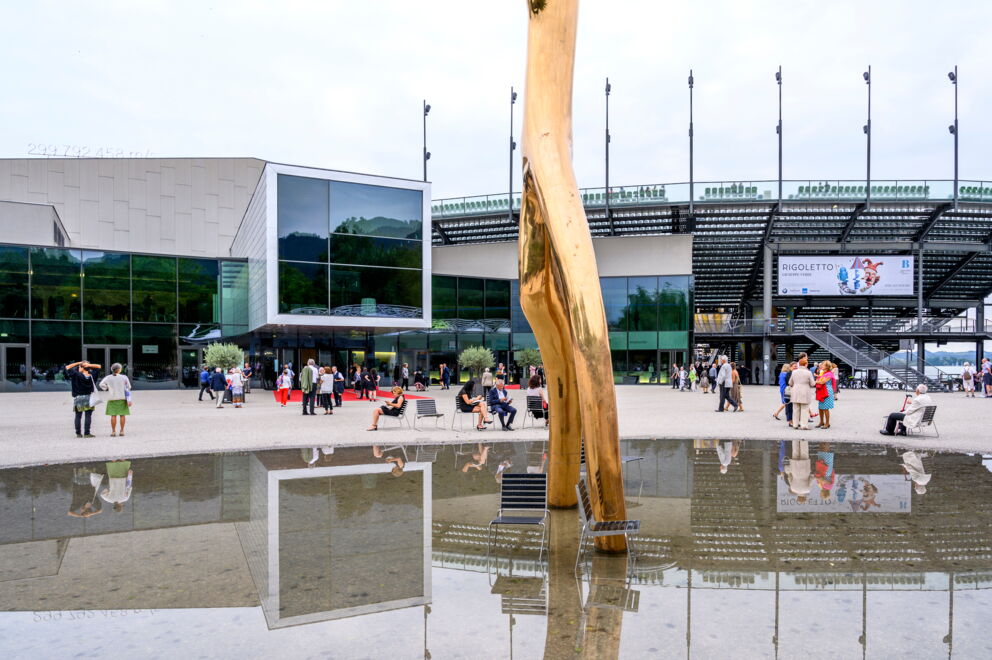 Menschen vor moderner Glasfassade mit Kunstinstallation und Wasserspiegelung.