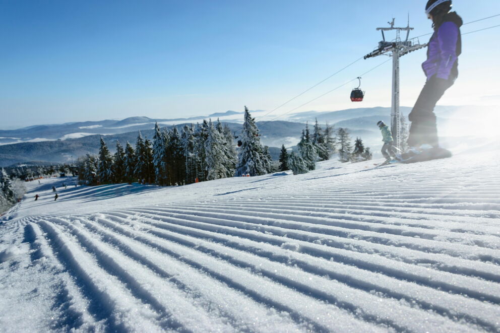 Skiers on a freshly groomed slope with a snow-covered mountain panorama and chairlift.