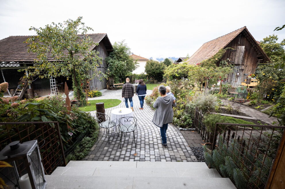Family strolls through charming garden with rustic buildings and flowering plants.