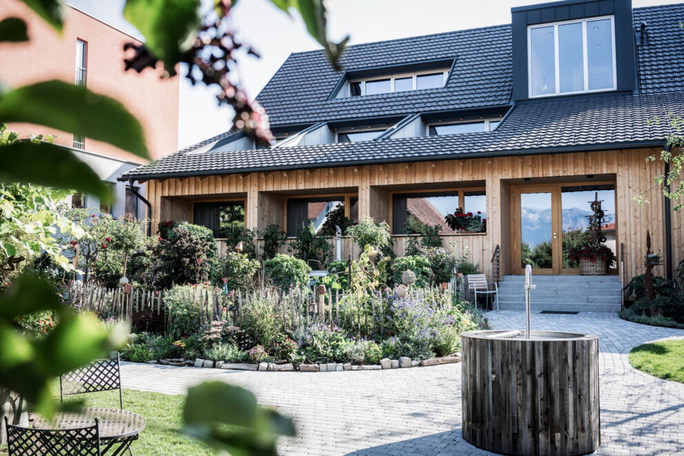 A lush, flowering garden in front of a charming wooden building with patio windows.