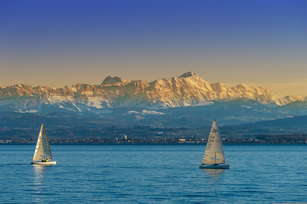 Two sailing boats on a lake with snow-capped mountains in the background.