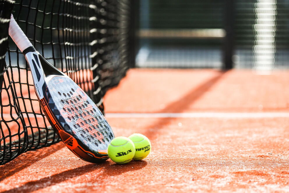 Padel-Schläger und Bälle auf rotem Platzboden neben Netz im Sonnenlicht.