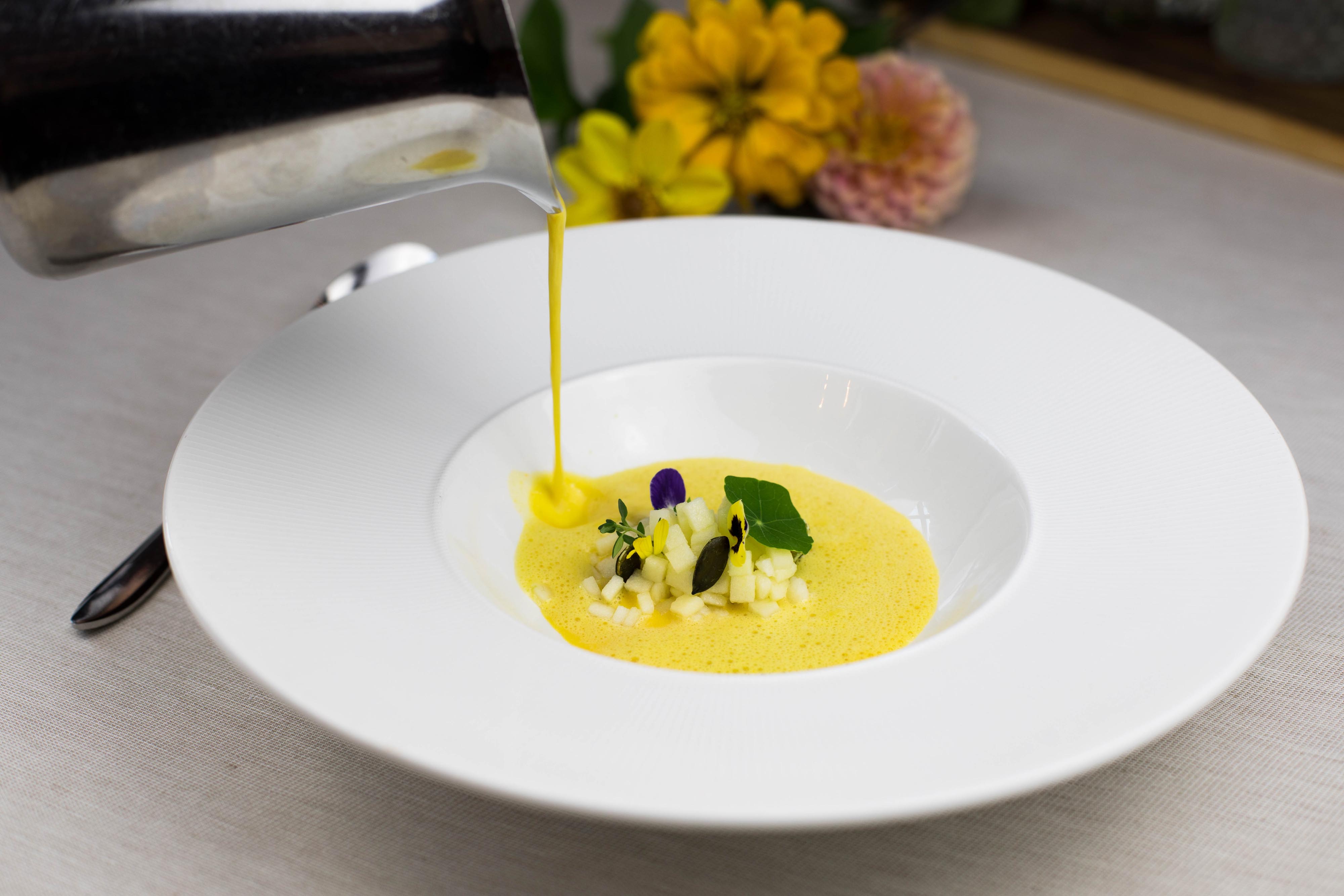 Gelbe Suppe wird kunstvoll in einen Teller mit Blumen- und Kräutergarnitur gegossen.